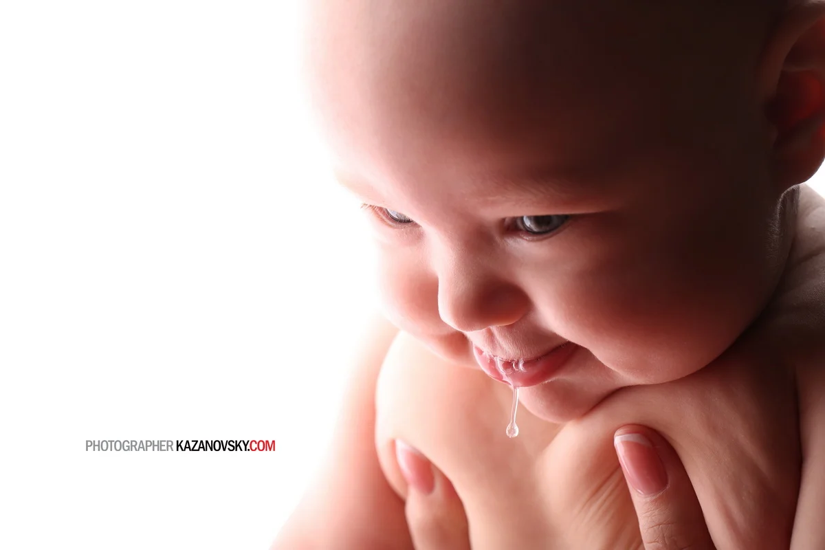 Close-up of a baby with a droplet of drool hanging from their mouth, smiling with a hand supporting their chest, and light background.
