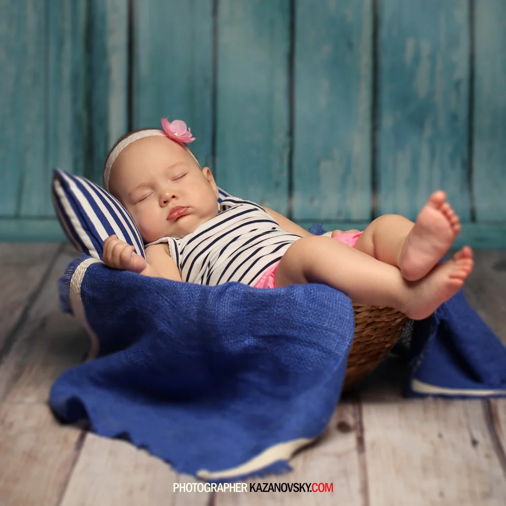 A sleeping baby girl lying on a small pillow in a wicker basket with a blue blanket, wearing a striped dress and a pink flower headband, against a teal wooden background.
