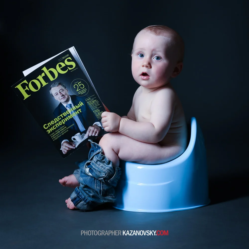 A baby sitting on a toilet holding a Forbes magazine, with a surprised expression, against a dark background.