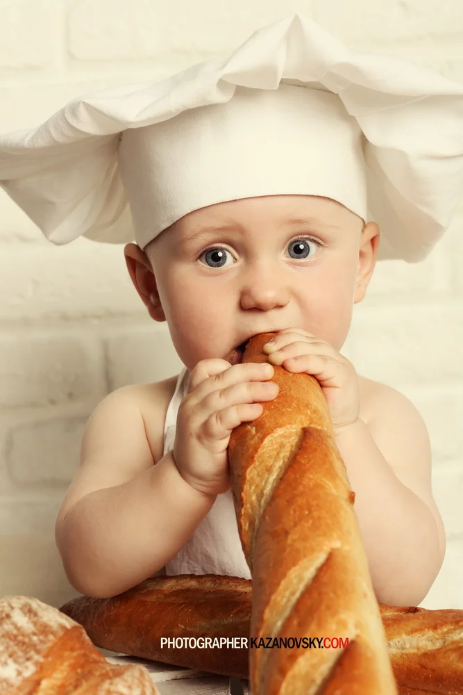 A young child wearing a white chef hat and apron is holding and biting into a baguette, with a few other baguettes visible in the foreground.