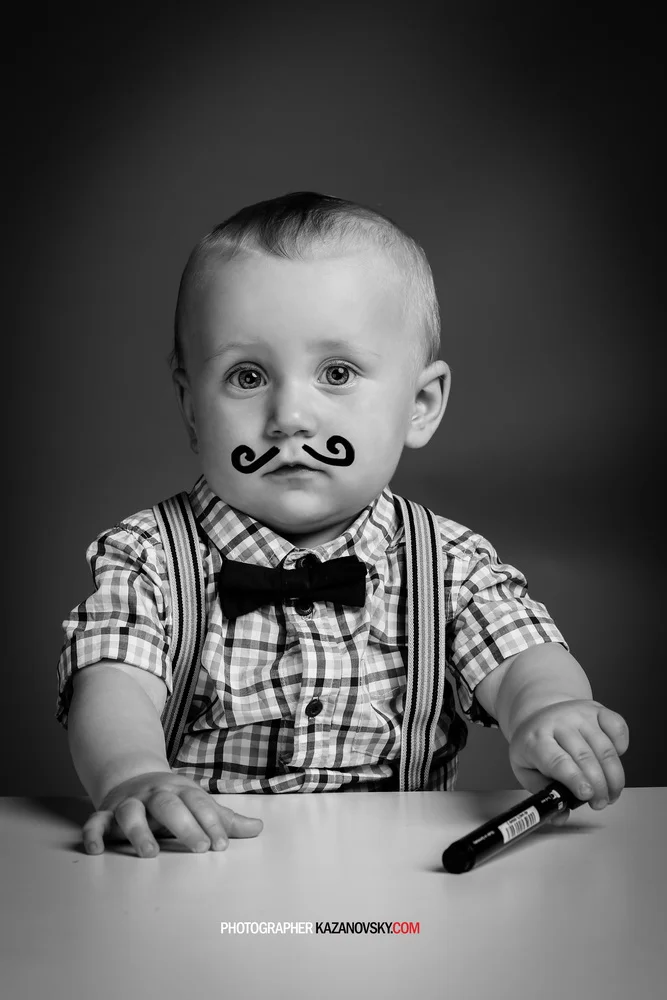 Black and white photo of a young boy with a drawn mustache on his face, wearing a checkered shirt, suspenders, and a bow tie, sitting at a table with a marker in his hand against a plain background.