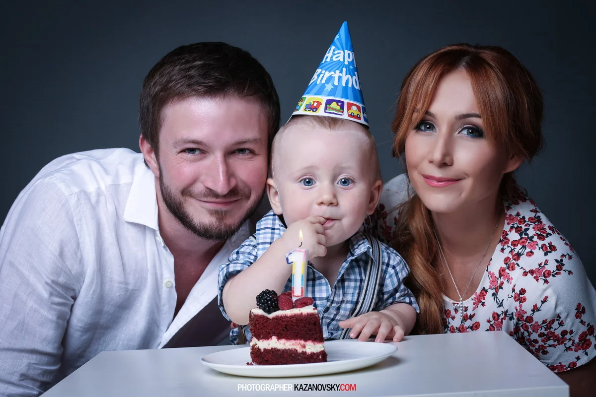 A young boy wearing a birthday hat celebrating his first birthday with a slice of cake, flanked by a man and woman, possibly his parents, all smiling at the camera.