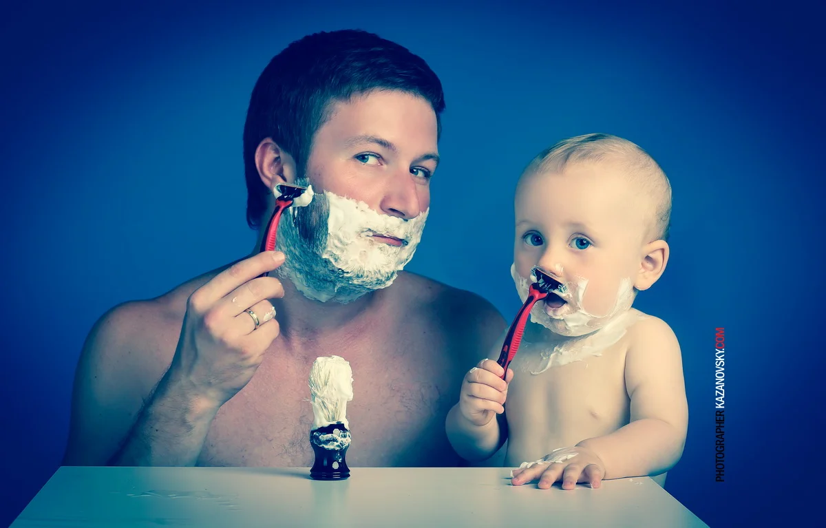 A man and a baby, both shirtless, are shaving with razors and have shaving cream on their faces, holding razors with cream on their faces, against a blue background.