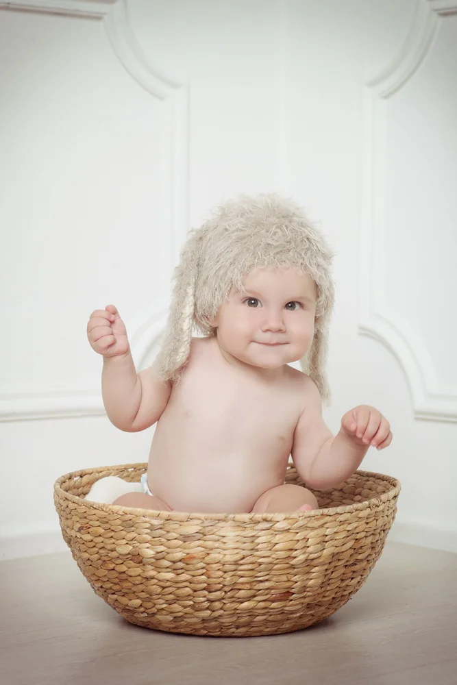 A baby sitting in a wicker basket, wearing a fluffy beige hat with ear flaps, in a white room.