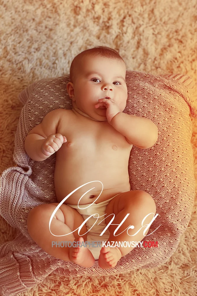 A baby with light skin and short hair lying on a plush, textured pink blanket, with one hand near their mouth and the other clenched in a fist, looking at the camera.