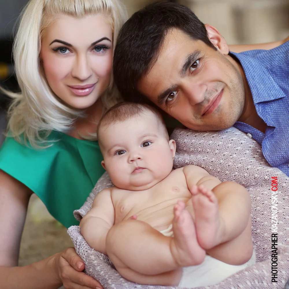 A young family with a mother, father, and baby lying close together, smiling at the camera.