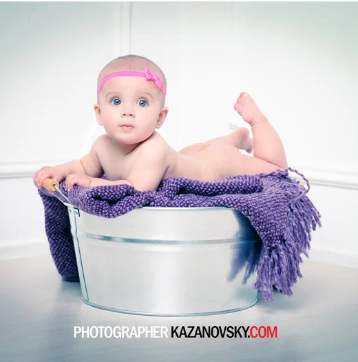 Baby lying on a purple blanket inside a metal tub, wearing a pink headband, with a surprised expression.