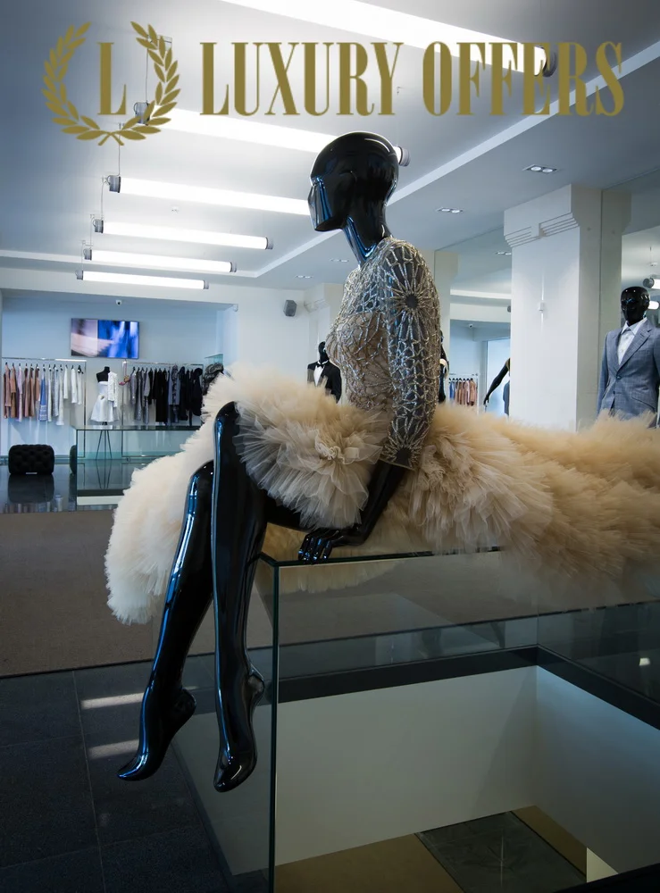 Display mannequin in a gold and black lace dress with a fluffy beige tulle skirt, sitting on a glass display case in a high-end clothing store, with a sign overhead reading 'Luxury Offers'.