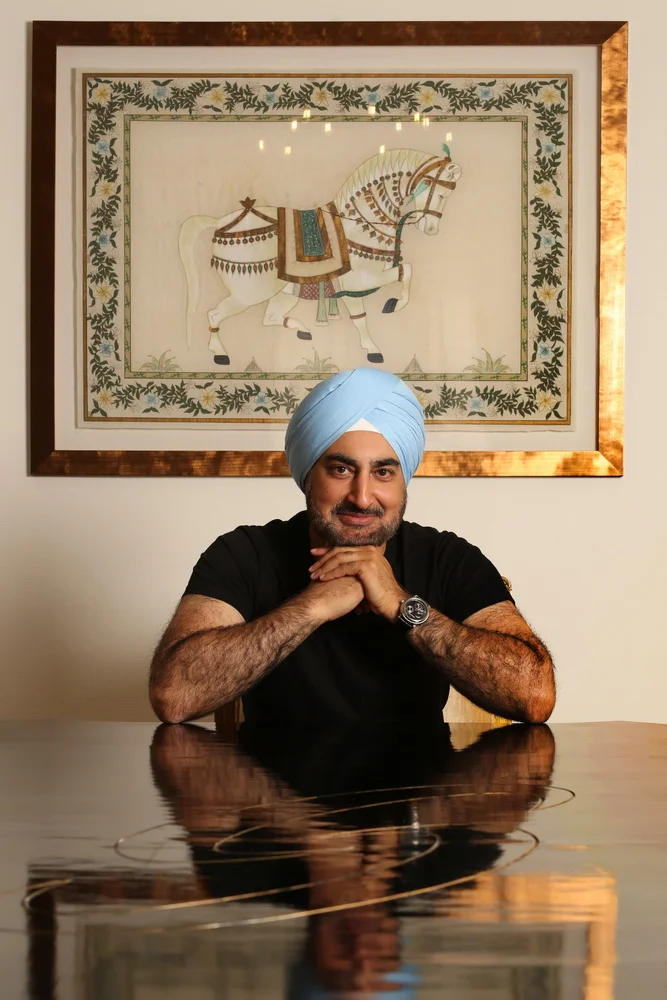 A man wearing a light blue turban and a black t-shirt, smiling with hands clasped, seated at a reflective wooden table, with a framed artwork of a decorated white horse hanging on the wall behind him.