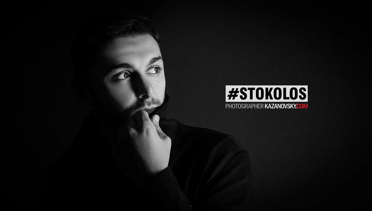 Black and white portrait of a young man looking to the side with his hand on his chin, wearing a dark jacket, and a dark background, with text overlay '#STOKOLOS PHOTOGRAPHER KAZANOVSKY.COM'.