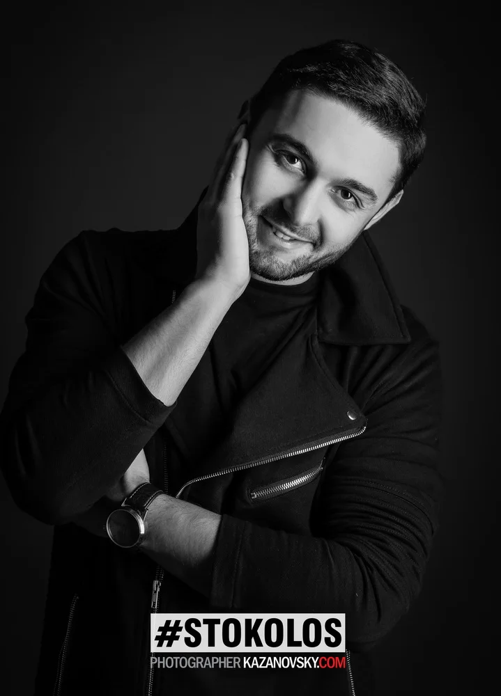 Black and white portrait of a smiling man with short hair, wearing a dark jacket and a watch, holding his face with one hand, against a dark background. Text overlay includes hashtag #STOKOLOS and a website link.