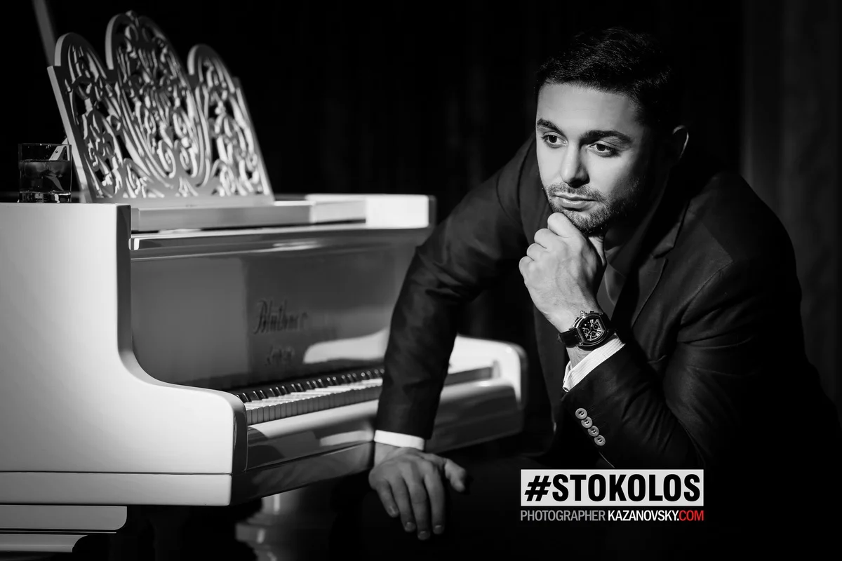 A man in a suit leaning on a white grand piano in a black and white photo, with a glass of water on top of the piano, and an ornate music stand. The man has short hair, a beard, and appears to be posing thoughtfully, wearing a watch on his left wrist