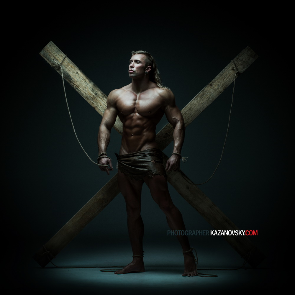 A muscular man with long hair is posed in front of a large wooden cross, simulating the crucifixion, with minimal clothing and dark background.