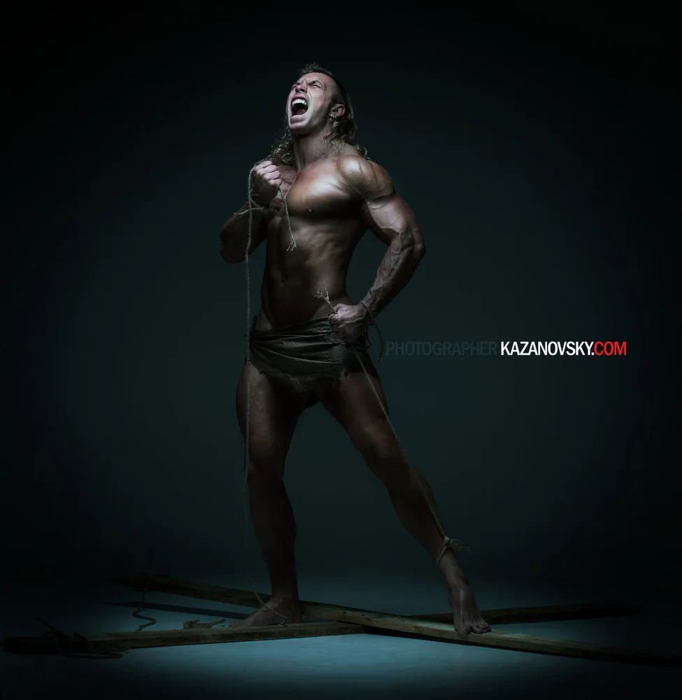 A muscular man with long hair, shirtless, standing on a dark studio floor with a dark background. He is holding a rope tied to his chest, with one fist clenched, showing an intense expression.