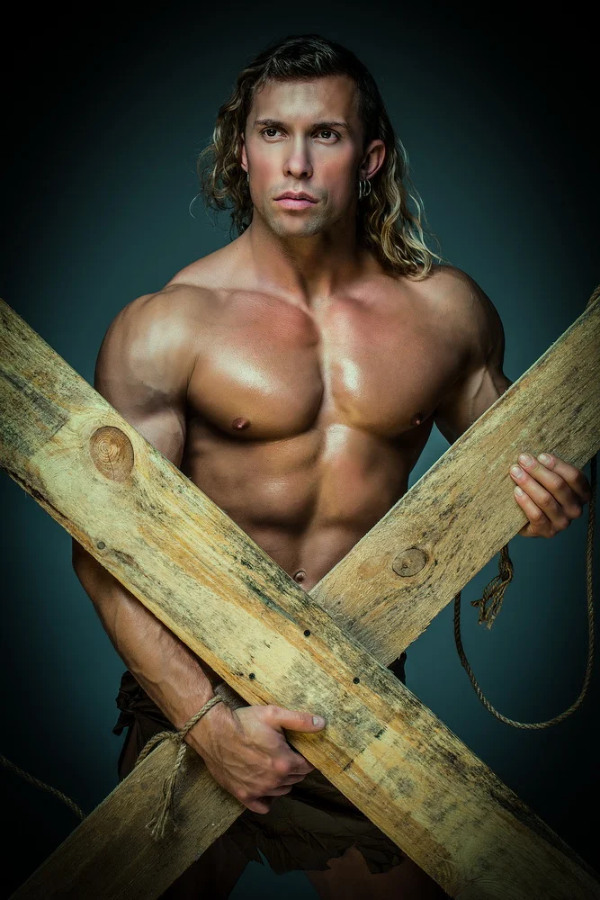 Muscular shirtless man holding a wooden cross, against a dark background.