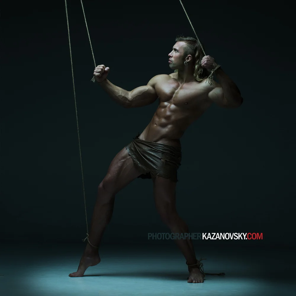 A muscular man with long hair and light skin posing shirtless with ropes tied around his wrists and ankles in a dark studio setting.