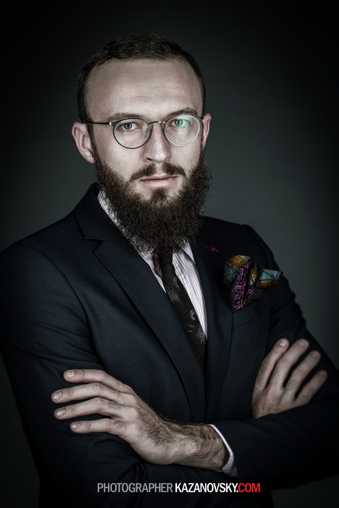 Man with glasses and a beard wearing a dark suit and tie, standing with crossed arms against a dark background.