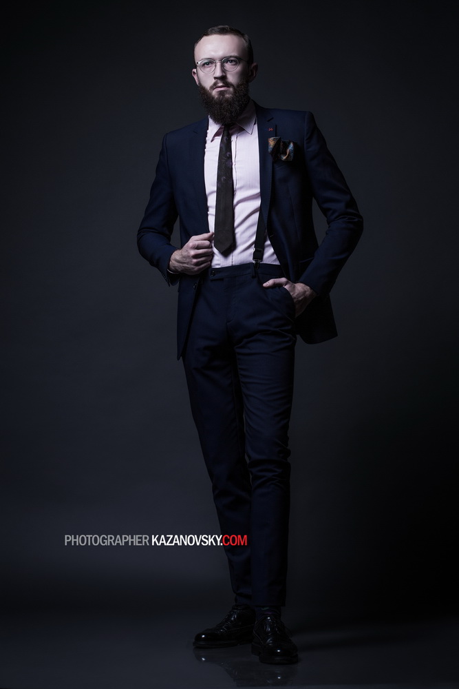 A man with a beard and glasses wearing a navy suit, pink shirt, black tie, and suspenders stands against a dark background. He has one hand in his pocket and the other adjusting his tie, and is looking directly at the camera.
