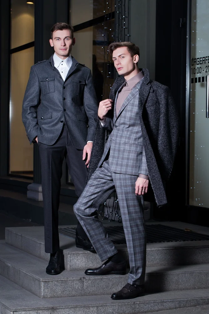 Two young men dressed in stylish formal suits stand on steps outside a building at night, one with hand in pocket, the other with hand on coat lapel.