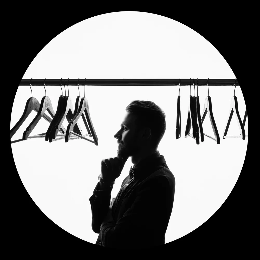 Silhouette of a man with short hair and a beard, standing and looking thoughtfully at a row of empty clothes hangers on a rail against a backlit white background.