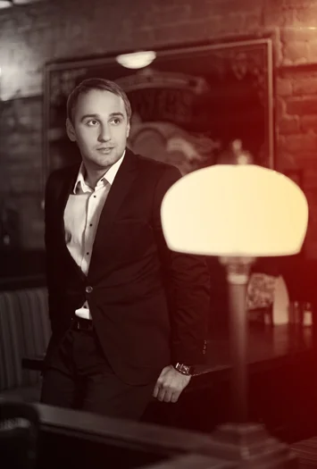 A young man in a suit standing in a dimly lit restaurant or bar, with a lamp in the foreground and brick walls in the background.