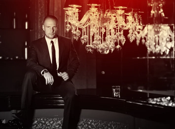 A man in a black suit sitting on a bench in a dimly lit room with a chandelier hanging above.