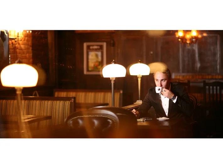 A man in a tuxedo sitting alone at a table in a dimly lit, vintage-style restaurant or bar, drinking from a white mug. The setting features warm lighting, lamps, wooden walls, and leather chairs.