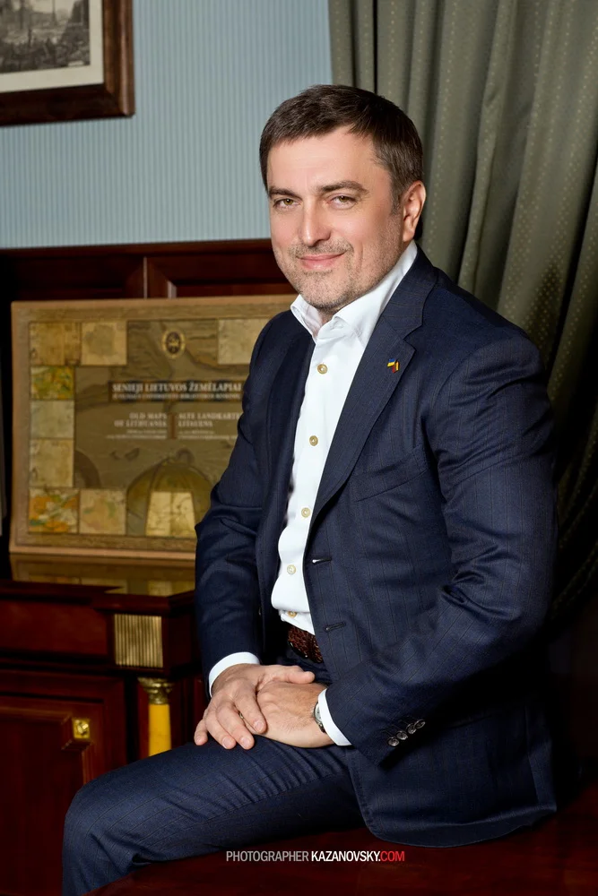 A man with brown hair and a beard, wearing a navy suit and white shirt, sitting on a wooden surface indoors, with a framed picture on the wall behind him and a wooden cabinet with a decorative plaque. He has his hands resting on his knee and is looki