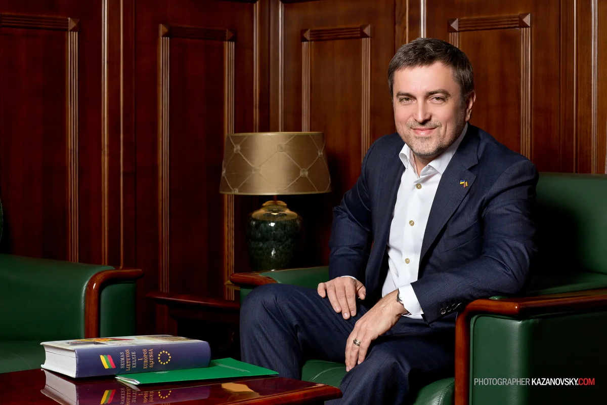 A man in a dark suit and white shirt sitting on a green wooden bench in a wood-paneled room, with a table in front of him holding an open book. The room features a lamp on a side table and wooden panel walls.