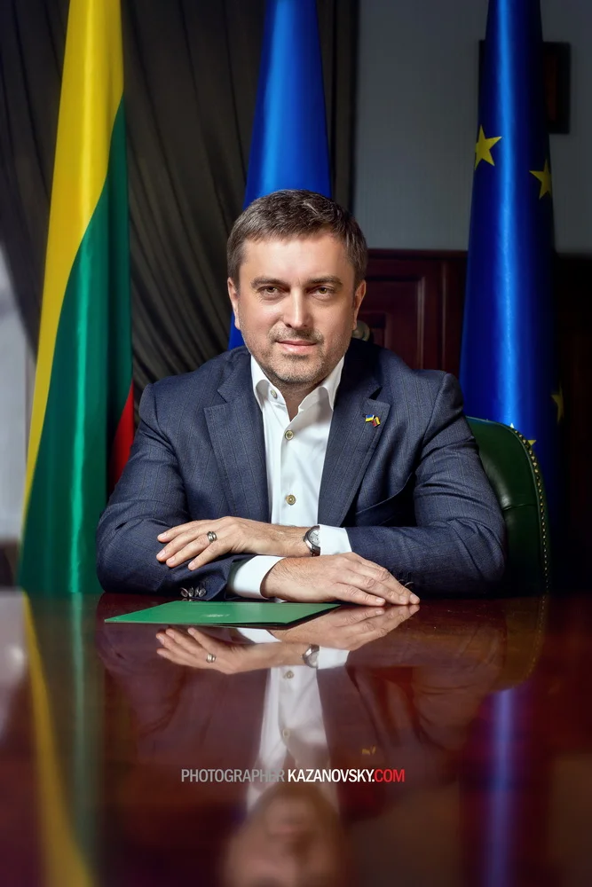 A man in a dark suit and white shirt sitting at a reflective wooden table, with flags behind him including a Lithuanian flag, a European Union flag, and a blue flag, in an official setting.
