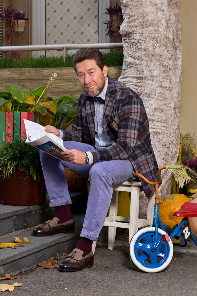 A man with dark hair and a beard sits outdoors on a small wooden stool, reading a magazine. He's dressed in a plaid blazer, light blue pants, and brown dress shoes. Behind him is a large tree and a planter with green and yellow plants. To his right i