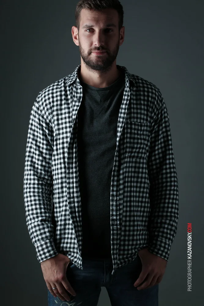 A man with dark hair and a beard wearing a black and white checkered shirt over a dark t-shirt, standing against a gray background.