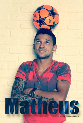 Man with tattoos and watch smiling with arms crossed, balancing an orange soccer ball with blue stars and logos on his head in front of a white brick wall. The word "Matheus" is written at the bottom.