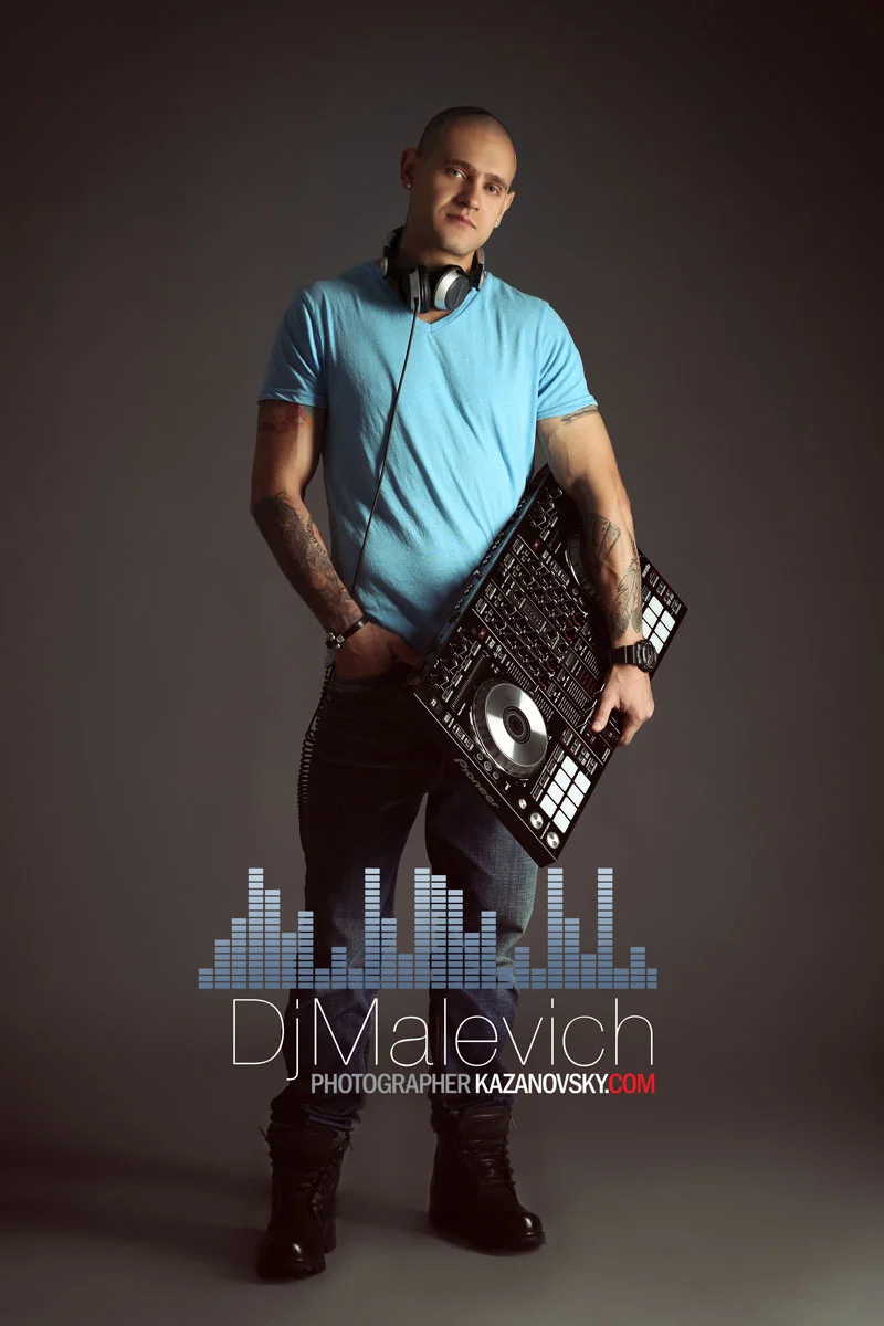 Portrait of a DJ holding DJ equipment, wearing a blue t-shirt and headphones around his neck, standing in front of a plain dark background with a sound wave graphic and artist information at the bottom.