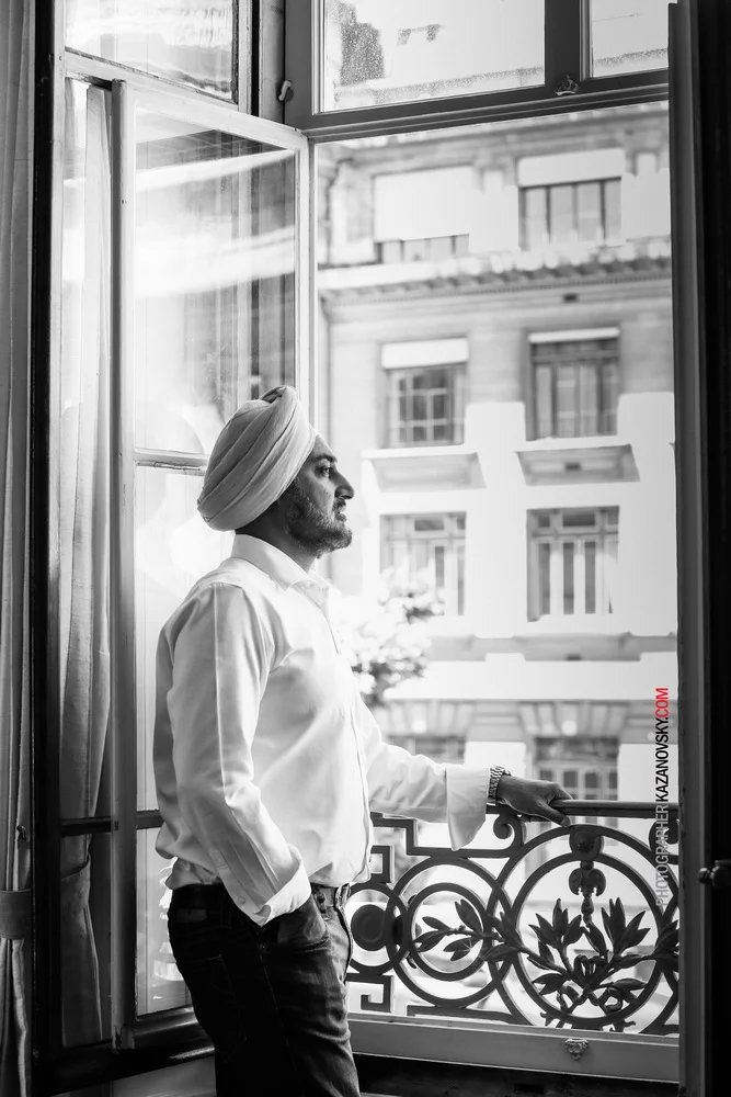 A man wearing a turban and white shirt standing by an open window, looking outside, in a black and white photo.
