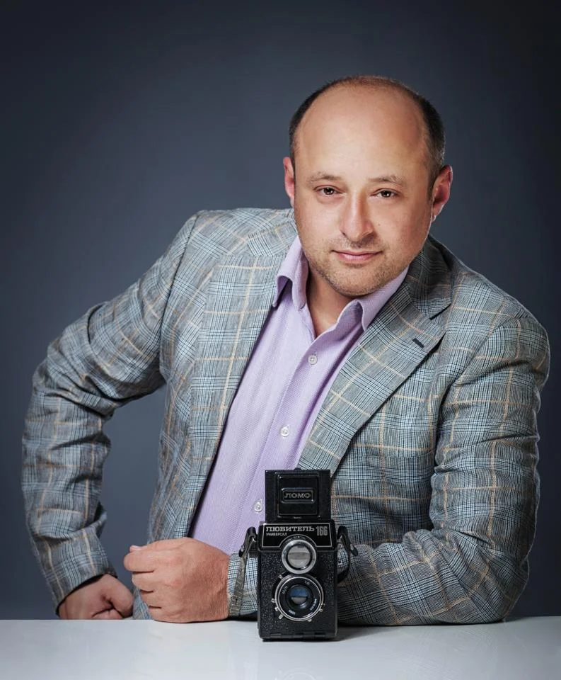 A man in a patterned blazer and light purple shirt poses with a vintage twin-lens reflex camera on a white surface against a dark background.