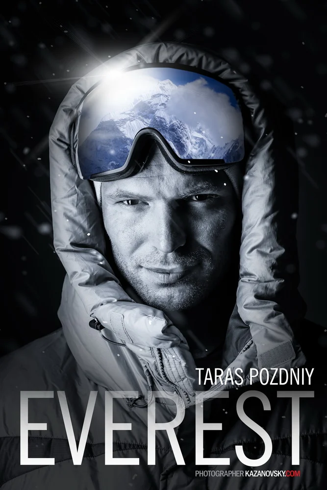 Close-up of a man in winter gear with a reflective mountaineering goggles showing an icy mountain landscape. Snow falls around him, and he wears a padded jacket with a hood.