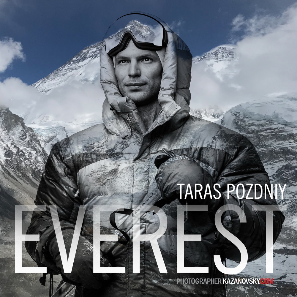 Black and white photo of a man in outdoor winter gear with mountain scenery in the background, holding a camera near his chest.