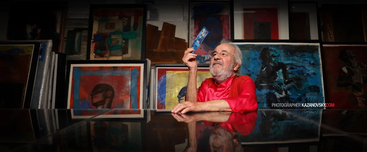 An older man with gray hair and a beard, wearing a red shirt, sitting at a table in an art studio or gallery surrounded by colorful abstract paintings, holding a small art piece or photograph.