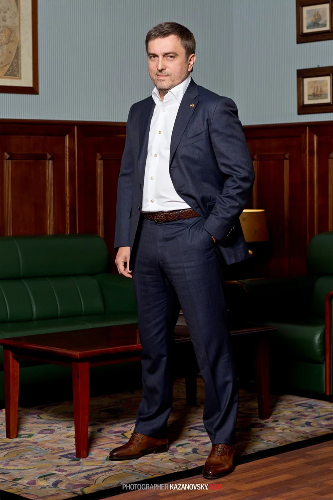 A man in a navy blue suit with a white dress shirt, standing in a room with wood-paneled walls, green leather furniture, and framed pictures, looking at the camera with a confident expression.