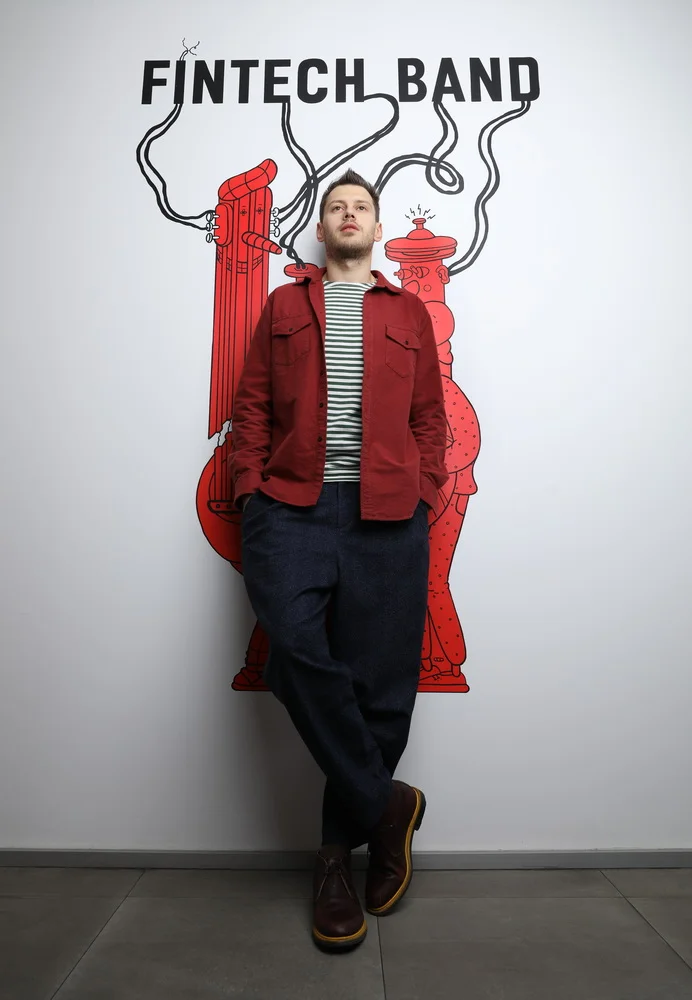 Man standing in front of a wall with a red artwork and the words 'Fintech Band' at the top. He is wearing a red jacket, striped shirt, dark pants, and brown boots.