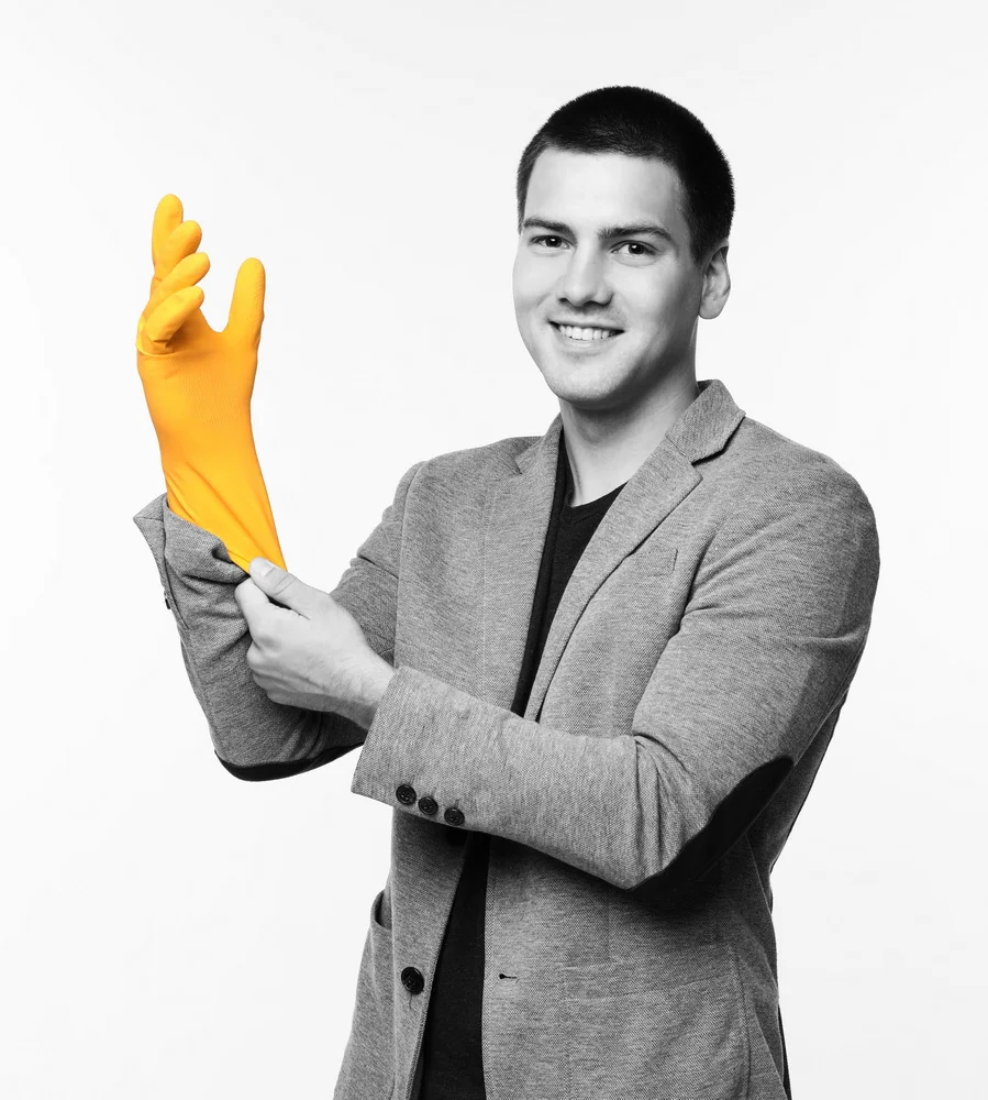 A young man in a gray blazer smiling and wearing a yellow rubber glove on his left hand, holding his arm as if putting it on, against a plain white background.