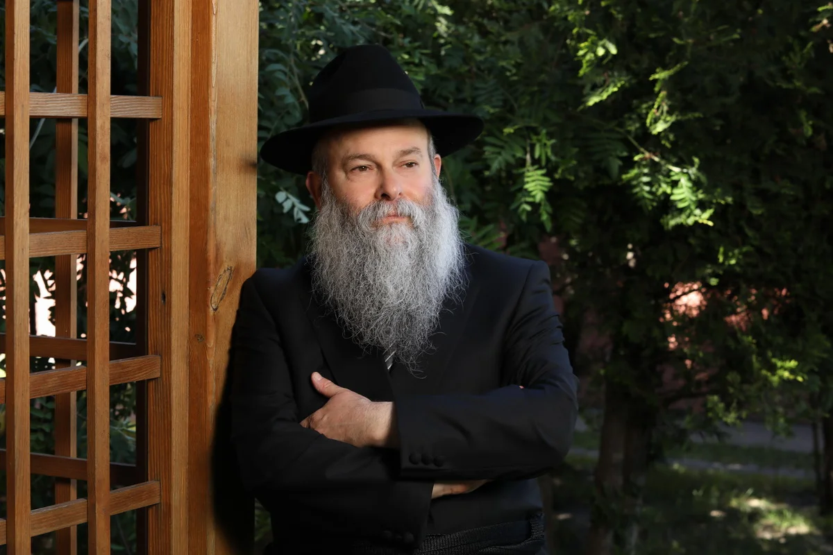 A man with a long grey beard wearing a black suit and black hat, standing outdoors with arms crossed near a wooden lattice and green foliage in the background.