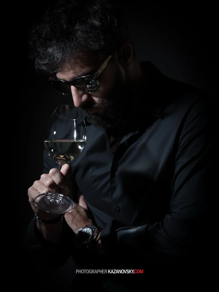 A man with dark curly hair, glasses, and a beard holding a wine glass up to his face, looking at it. He is wearing a black shirt and a watch, against a dark background.