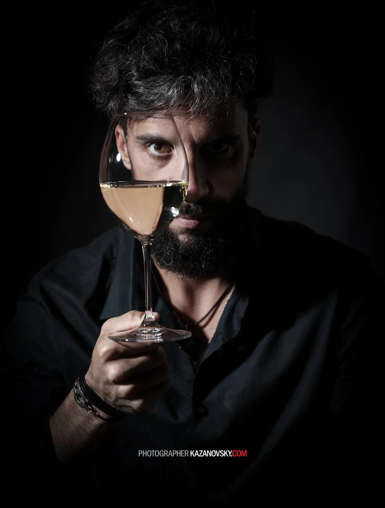 A man with dark hair and a beard holding a wine glass filled with white wine, with part of his face visible through the glass.