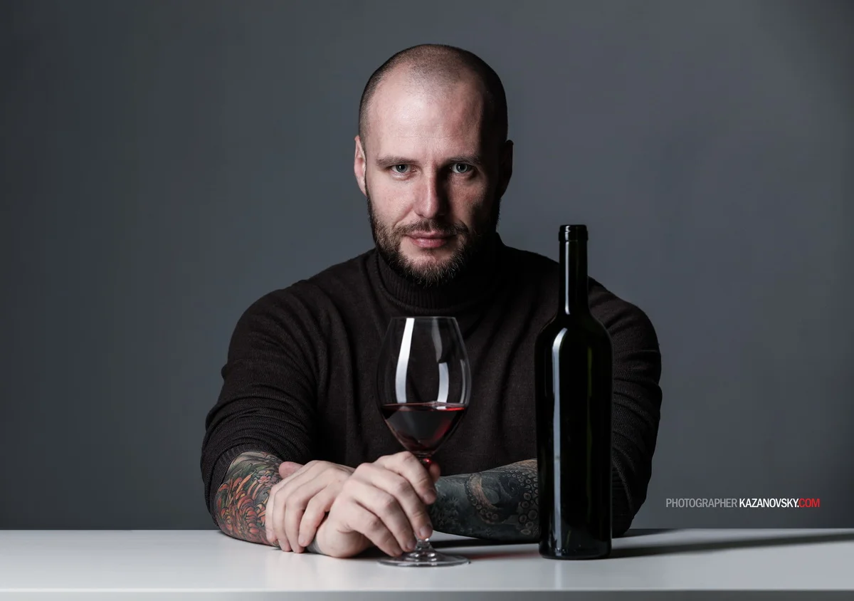 A man with a shaved head, beard, and colorful tattooed arms sitting at a table with a glass of red wine and a wine bottle in front of him against a gray background.