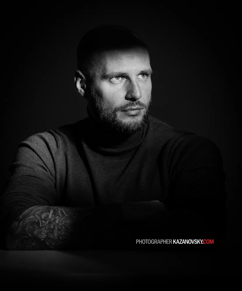 Black and white portrait of a man with a beard and short hair, wearing a turtleneck sweater, looking to the side with a serious expression, with a tattooed arm resting on a surface, against a dark background.