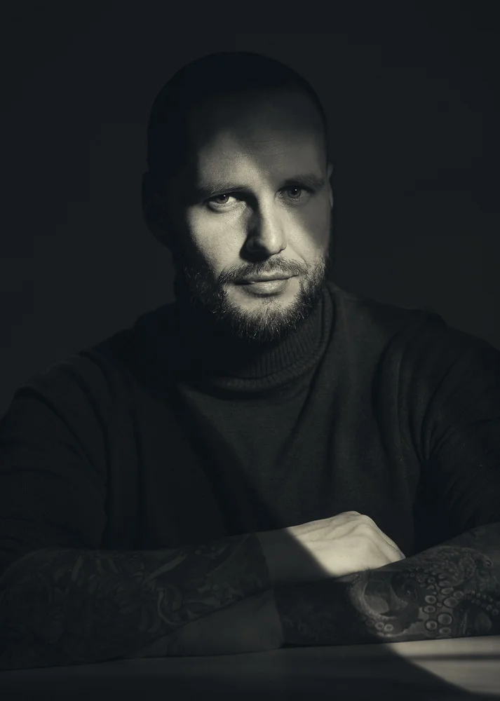 A black and white portrait of a man with a beard, short hair, and tattoos on his arms, wearing a black turtleneck sweater, looking directly at the camera with a serious expression.
