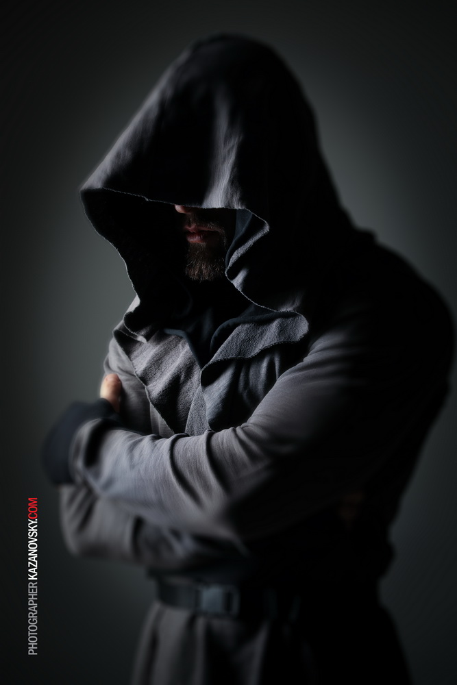 A person in a dark hoodie with the hood up, partially covering their face, standing with arms crossed. The background is dark and the lighting highlights the contours of the person's clothing and face.