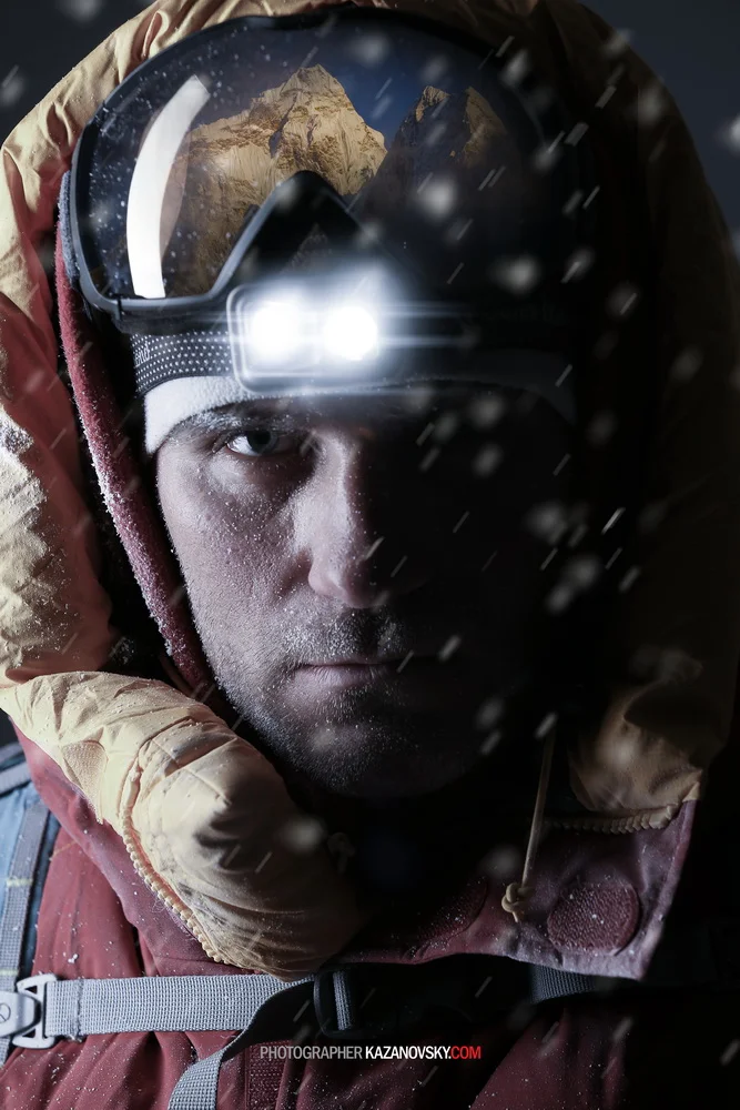 A climber wearing a ski mask and headlamp, with snow on his face, dressed in a brown and red winter jacket, with a mountainous landscape reflected in his goggles.